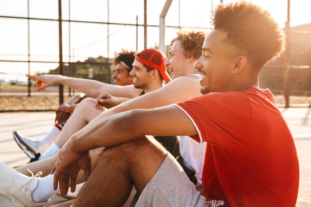 Photo of sporty athletic guys sitting at basketball playground outdoor and watching game during summer sunny day - Photo, image