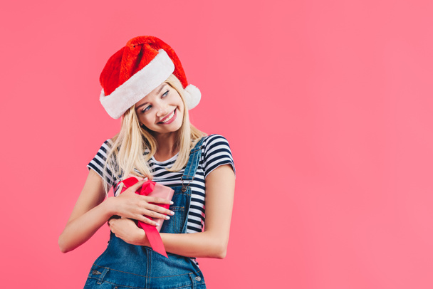 portrait of young happy woman in santa claus hat holding gift isolated on pink - Foto, Imagen