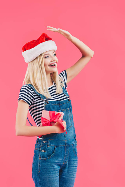 young cheerful woman in santa claus hat holding gift isolated on pink - Photo, Image