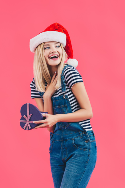 portrait of smiling woman in santa claus hat with heart shaped gift isolated on pink - 写真・画像