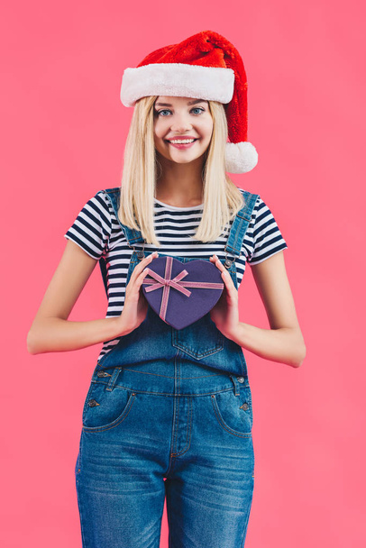 portrait of smiling woman in santa claus hat with heart shaped gift isolated on pink - Foto, imagen