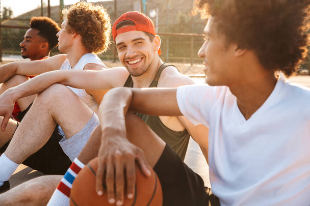 Group of excited multiethnic friends basketball players sitting at the sports ground, talking - Zdjęcie, obraz