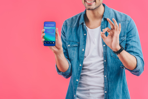 partial view of young man doing ok sign and showing smartphone with booking isolated on pink - Foto, Imagen