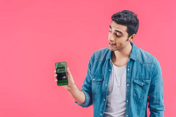 smiling young man showing smartphone with booking isolated on pink - Foto, Imagen