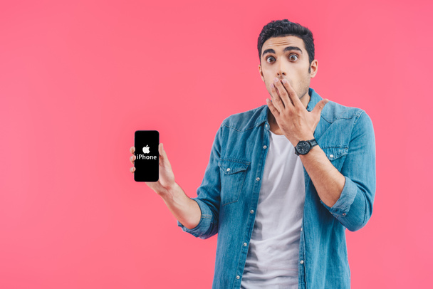 shocked young man covering mouth by hand and showing iphone smartphone isolated on pink - Fotografie, Obrázek