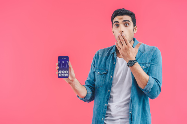 chocado jovem cobrindo a boca com a mão e mostrando smartphone com aplicação médica isolada em rosa
 - Foto, Imagem