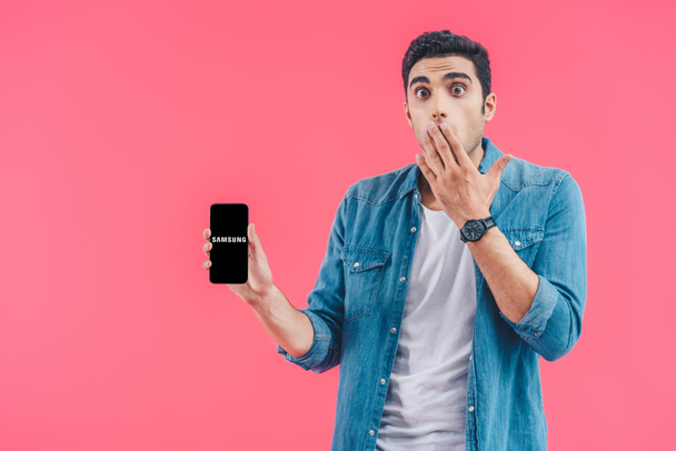 shocked young man covering mouth by hand and showing samsung smartphoneisolated on pink - Foto, Imagem