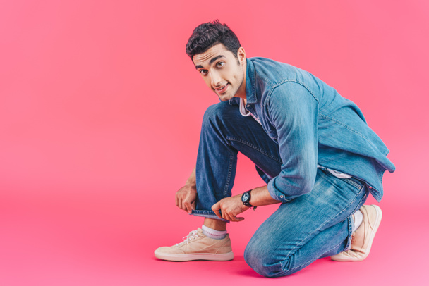 young stylish man sitting and adjusting jeans isolated on pink - Fotó, kép