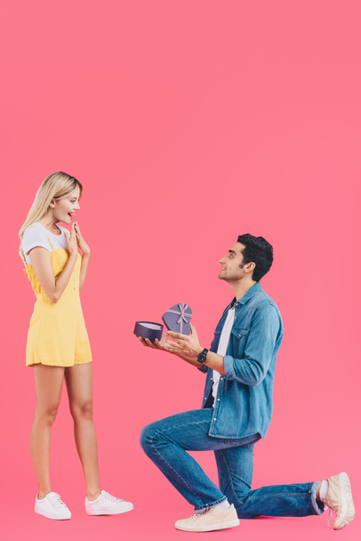 Zijaanzicht van de mens op gebogen knie doet huwelijksaanzoek om verrast vriendin gebaren door handen geïsoleerd op roze - Foto, afbeelding
