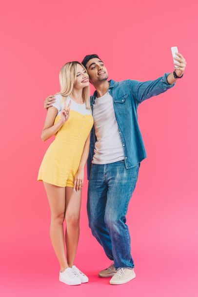 красивий молодий чоловік бере селфі з дівчиною робить знак миру на смартфоні ізольовано на рожевому
  - Фото, зображення