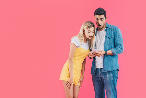 Šokovaný mladý muž dívá na smartphone, zatímco jeho přítelkyně, stojící poblíž izolovaných na růžové - Fotografie, Obrázek