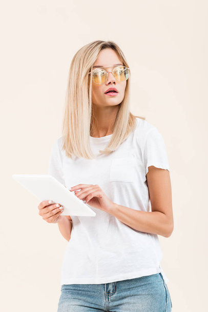 attractive young woman in eyeglasses looking away and holding digital tablet isolated on beige - Fotoğraf, Görsel