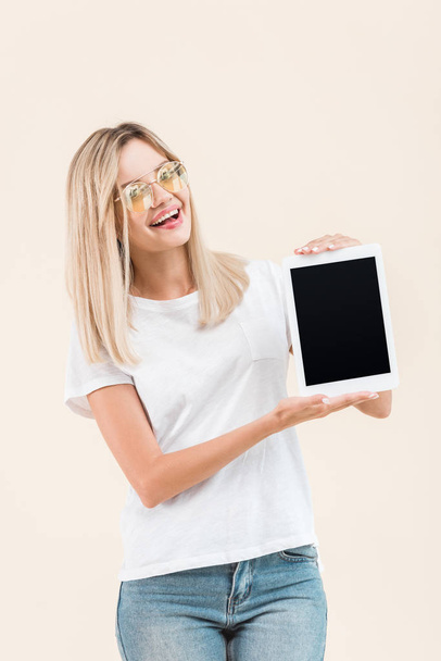 cheerful young woman in stylish eyeglasses showing digital tablet with blank screen isolated on beige - Фото, изображение