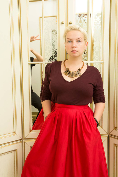 young blonde in a red sweater, burgundy skirt and ethnic ornaments next to a mirror - Photo, image