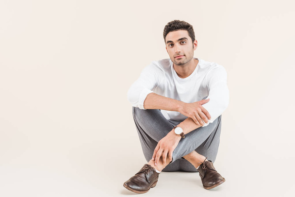 full length view of handsome young man sitting and looking at camera isolated on beige - Foto, Bild