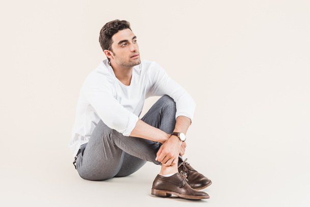 full length view of pensive young man sitting and looking away isolated on beige - Photo, Image