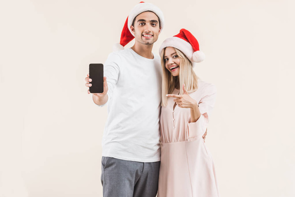 glückliches junges Paar in Weihnachtsmannmützen, Smartphone mit leerem Bildschirm in der Hand und lächelnd in die beige isolierte Kamera  - Foto, Bild