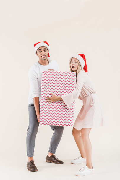 young couple in santa hats holding big christmas present and looking at camera isolated on beige - Fotografie, Obrázek