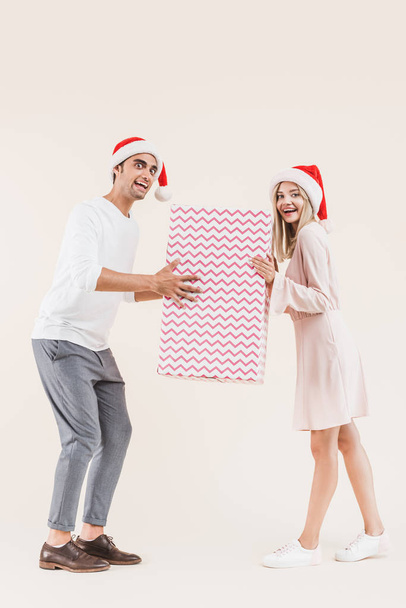happy young couple in santa hats holding big christmas gift and smiling at camera isolated on beige  - Fotó, kép