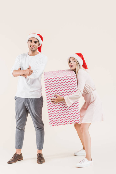 happy young man in santa hat showing thumb up and smiling at camera while woman holding big christmas gift isolated on beige - Фото, изображение