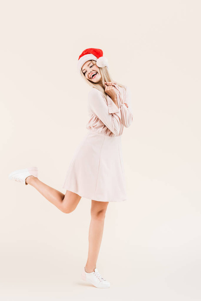 full length view of beautiful happy young woman in dress and santa hat isolated on beige - Foto, immagini