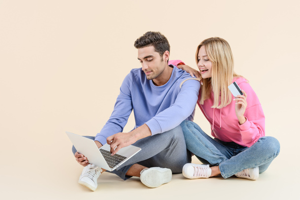 Lächelndes junges Paar mit Kreditkarte mit Laptop isoliert auf beige - Foto, Bild