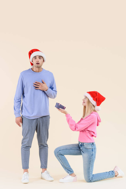 smiling young woman kneeling and presenting gift box to shocked young man in santa hat isolated on beige - Foto, Imagem
