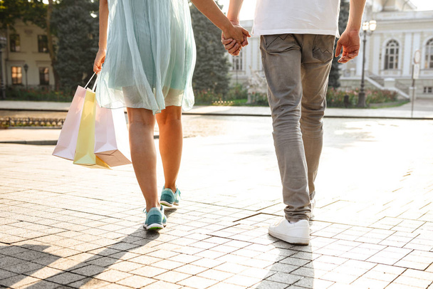 Cropped photo of young cute loving couple walking outdoors while holding hands of each other and shopping bags. - Фото, изображение