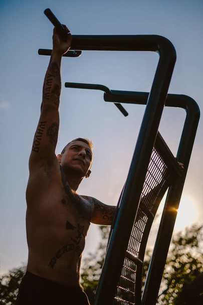 Shirtless macho man with tattoos climbing on outdoor gym pull up bar - Foto, imagen