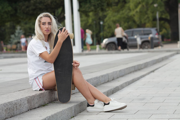 Casual blond female in denim shorts posing on stairs with longboard. - Foto, Bild