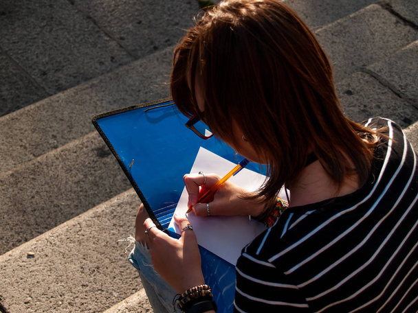 Μια Έφηβη γυναίκα γράφοντας με μια μάνδρα σε ένα προφίλ σε ένα φάκελο που κάθονται στα σκαλοπάτια σε μια πανεπιστημιούπολη Πανεπιστήμιο - Φωτογραφία, εικόνα