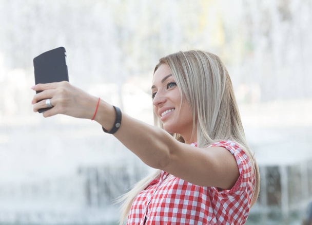 Carefree and happy, sunny summer mood. Charming young lady is making selfie on a camera. She is wearing formal wear, smiling, while on a walk in town outdoors. - Valokuva, kuva