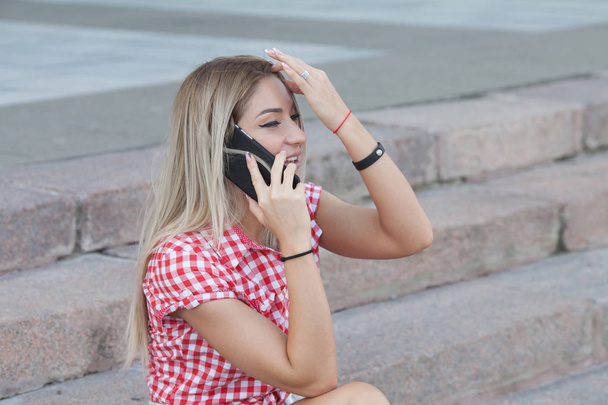 Close up of smiling attractive girl with long hair talking on mobile phone while standing outdoors on a city street. - Фото, изображение