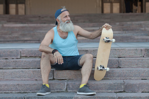 trendy old bearded man  with skateboard in town, relaxing on stairs - Foto, Bild