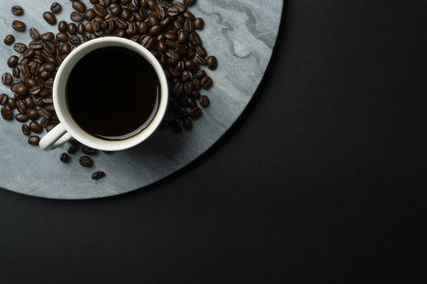 top view of a cup of coffee and a pile of coffee beans on the round marble tray, black background - Foto, Imagem