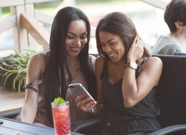 Multiethnic women read notification with positive news on mobile phone, spend free time in terrace cafe, embrace each other, have happy expressions. Two diveristy females choose something in internet - Photo, Image