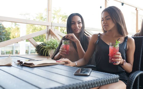 female couple have postive expressions, sit close to each other at cafeteria, smile joyfully, enjoy tasty desserts in outdoor cafeteria. Multiethnic lesbians talk to each other. Love concept. - Photo, Image