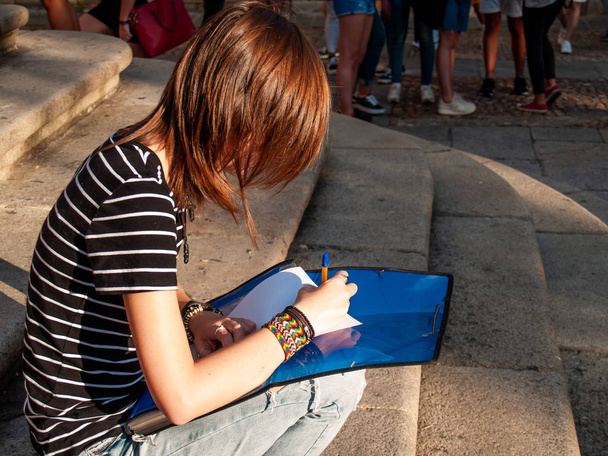 Подросток, пишущая ручкой в фолио на папке, сидящей на лестнице университетского кампуса
 - Фото, изображение