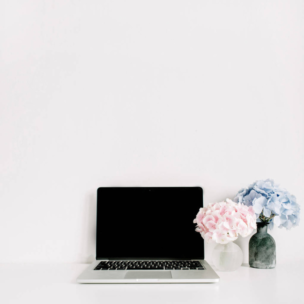Prázdná obrazovka přenosného počítače a růžové a modré hortenzie kytice na bílém pozadí. Pracovní stůl prostor minimální domácí kancelář. - Fotografie, Obrázek