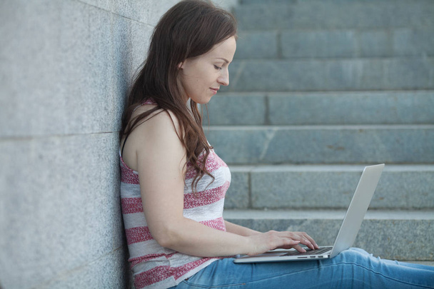 Portrait of a happy young woman sitting on the city stairs and using laptop computer outdoors - Фото, изображение