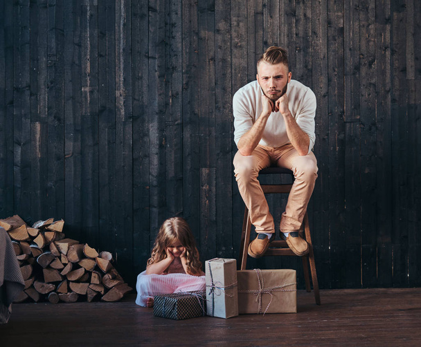 Verärgerter Vater und Tochter sitzen neben Kisten mit Geschenken im leeren Raum gegen Holzwand.  - Foto, Bild