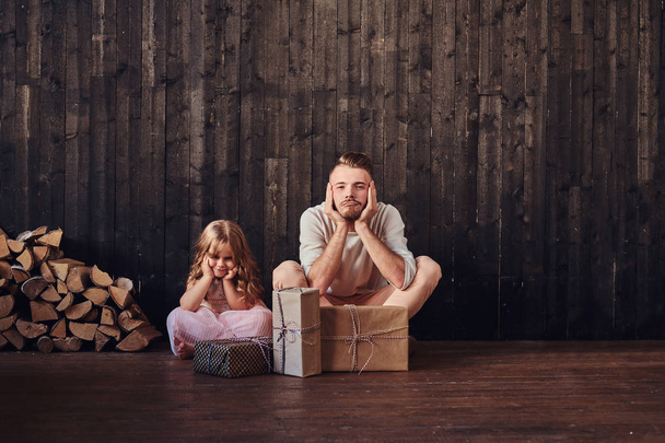 Vader en dochter zittend op de vloer in de buurt van vakken met giften in lege ruimte tegen een houten muur verstoren.  - Foto, afbeelding