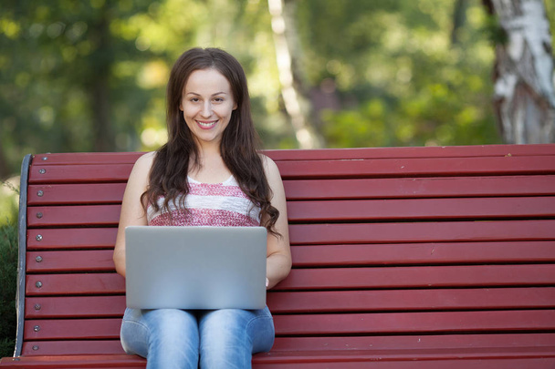 Piękna kaukaski kobieta siedzi na ławce w parku miejskiego z laptop podłączony do wi-fi. Przedsiębiorca działa na komputerze przenośnym siedząc na zewnątrz na niewyraźne tło miejskie. - Zdjęcie, obraz