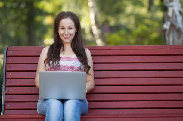 Beautiful caucasian woman is sitting at the bench in a city park with a laptop connected to wi-fi. Entrepreneur is working on a portable computer while sitting outdoors on a blurred urban background. - Foto, Bild