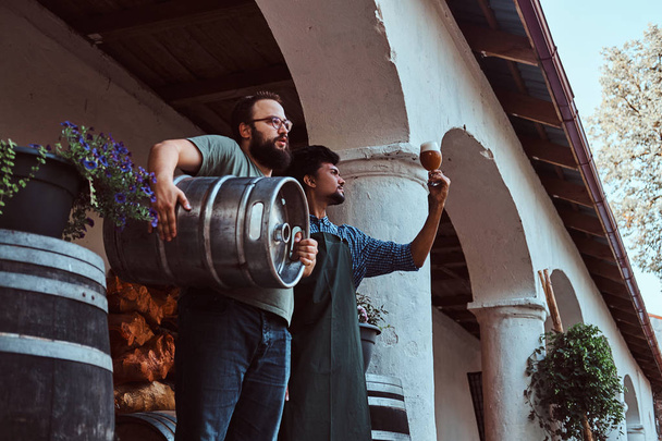 Dois cervejeiros em avental de pé ao ar livre verificando a qualidade da bebida fermentada na fábrica de cervejaria
. - Foto, Imagem