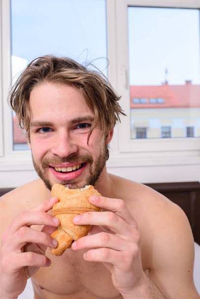Guy bites sweet dessert. Man bearded handsome guy eating breakfast in bed. Guy holds croissant bedroom hotel room window background. Enjoy every bite. Man eats croissant he likes bakery products - Photo, Image