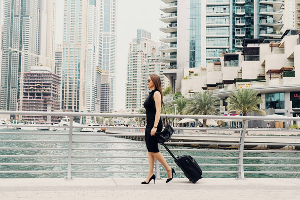 Fiatal modern magabiztos üzletasszony húz egy bőröndöt a Dubai Marine. Az új munkalehetőség a nagy városban. - Fotó, kép