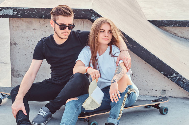 Trendy gekleed paar jonge schaatsers knuffelen elkaar zittend op skateboards in skatepark op zonnige dag. - Foto, afbeelding