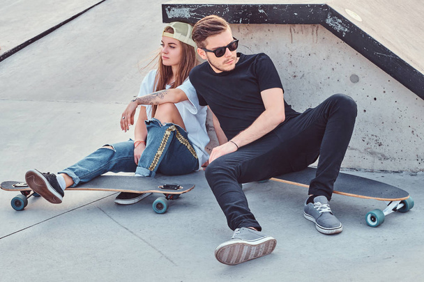 スケートパークでスケート ボードの上に座って若いスケーターのトレンディな服を着たカップル. - 写真・画像
