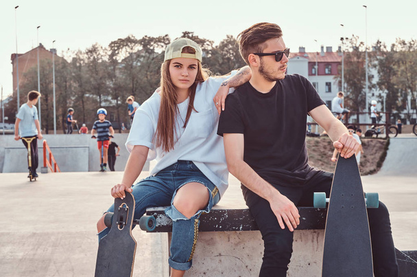 Young stylish couple of a trendy dressed skaters relaxing at skatepark. - Photo, Image